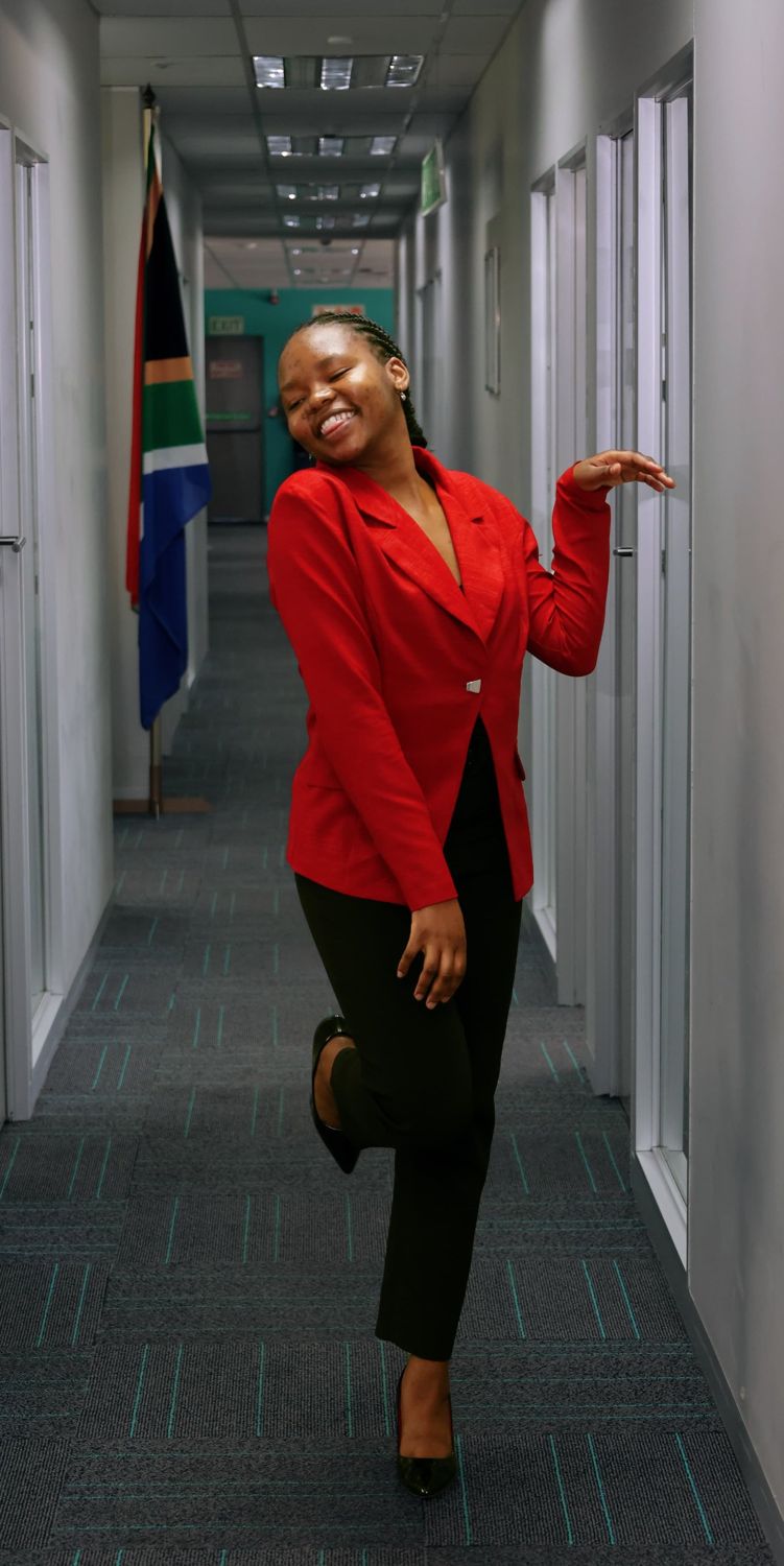 Woman in a red blazer posing playfully in an office corridor